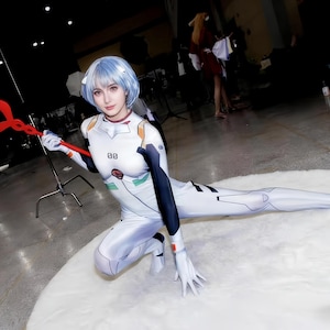 May include: A person in a white and black bodysuit with blue hair, holding a red spear. The costume has orange and green accents. The person is kneeling on a white, fluffy surface.