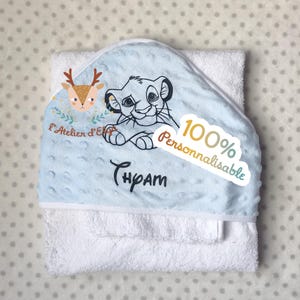 Op de afbeelding: Een witte babyhanddoek met capuchon met een lichtblauwe capuchon met een cartoon leeuwenontwerp. De capuchon heeft ook de tekst "Thpam" en een sticker met de tekst "100% Personnalisable". Er is ook een hertenillustratie aanwezig.