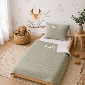 Può includere: Un set di biancheria da letto testurizzato color verde salvia su un letto in legno, composto da un cuscino e una coperta, entrambi ricamati con il nome Eloan e un cuore. La coperta presenta un lato inferiore in pile bianco e una superficie trapuntata.