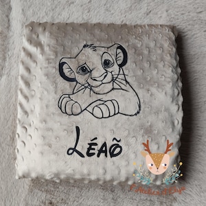 Puede incluir: Una manta cuadrada de bebé de color beige claro con un dibujo lineal negro de un cachorro de león. El nombre "LEÃO" está impreso en escritura negra debajo del león. La manta tiene una superficie texturizada y punteada.