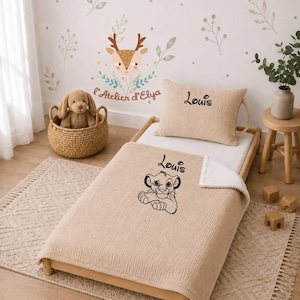 Puede incluir: Un conjunto de manta y almohada de color beige con textura sobre una estructura de cama de madera. La manta presenta un dibujo lineal negro de un cachorro de león y el nombre Louis. La almohada está bordada con el nombre Louis en negro.