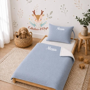 Peut inclure: Un ensemble de literie bleu clair texturé composé d'un oreiller rectangulaire et d'une couverture, tous deux ornés du prénom Alessio brodé en écriture blanche. La literie est disposée sur un cadre de lit en bois avec une peluche dans un panier.