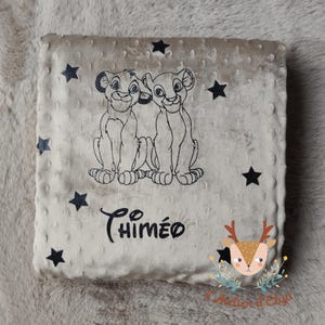 Puede incluir: Una manta de bebé beige con una superficie suave y texturizada. La manta presenta una ilustración en blanco y negro de dos cachorros de león, acentos de estrellas negras y el nombre "Chiméo" en escritura negra.