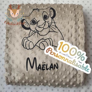 Op de afbeelding: Een beige babydeken met een zwarte omtrek van een leeuwenwelp. De deken heeft een getextureerd oppervlak met kleine, verhoogde stippen. De naam "Maëlan" is in het zwart gedrukt, met een sticker "100% Personnaliseerbaar".