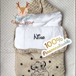 Könnte beinhalten: Eine beige Baby-Swaddle-Decke mit Kapuze und weißem Innenfutter. Die Decke zeigt eine Simba-Grafik, schwarze Sterne und den Namen "Nino" auf der Vorderseite. Der Text "100% Personnalisable" ist ebenfalls sichtbar.