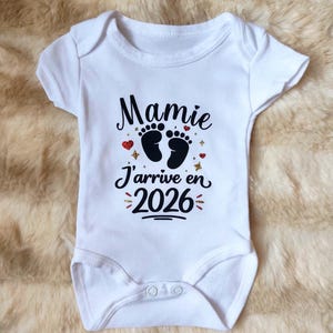 Può includere: Body da neonato bianco con la scritta francese "Mamie J'arrive en 2026" in nero. Il design include impronte di piedi di bambino, cuori rossi e stelle dorate. Il body è realizzato in tessuto morbido.