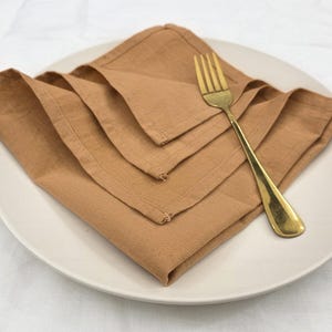 May include: A stack of folded, tan linen napkins on a white plate with a gold fork. The napkins are neatly arranged, showcasing their texture and color. The fork is positioned to the side, adding a touch of elegance to the dining setting.