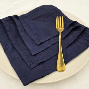 May include: A navy blue linen napkin set on a white plate with a gold fork. The napkins are folded and stacked, showcasing their texture. The fork is positioned to the right, adding a touch of elegance to the table setting.
