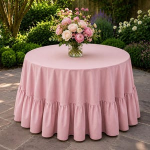 Pode incluir: Uma toalha de mesa redonda em rosa empoeirado com uma borda franzida, colocada sobre uma mesa circular. No centro da mesa, um vaso de vidro transparente contém um buquê de rosas cor-de-rosa e brancas com folhagem verde.