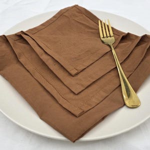 May include: A stack of four folded brown cloth napkins on a white plate. A gold-colored fork rests on the napkins. The napkins are arranged in a diamond shape, with the fork angled across them.