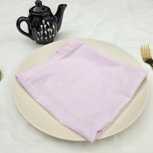 Puede incluir: Una mesa puesta con una cuchara y un tenedor dorados, un plato blanco con una servilleta rosa doblada y una tetera negra con detalles blancos. Los elementos están dispuestos sobre una superficie blanca.