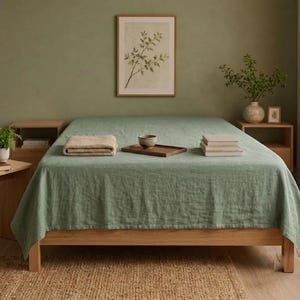 May include: A sage green linen bedspread draped over a wooden bed frame. On the bed, there is a folded beige blanket, a small dark wooden tray with a ceramic bowl, and a stack of books. The bed is flanked by two wooden nightstands.