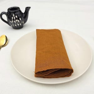 Op de afbeelding: Een tafelsetting met een witte plaat met een opgevouwen oranje servet, een gouden lepel en een gouden vork. Een kleine zwarte theepot met witte accenten is ook aanwezig. Op een wit oppervlak.