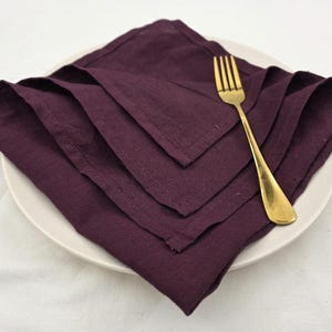 May include: A folded, deep purple linen napkin sits on a white plate, with a gold-coloured fork placed to the side. The napkin is square and has a textured appearance. The plate is round and simple, contrasting with the napkin.