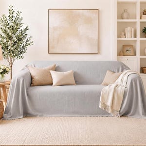 May include: A light gray sofa cover draped over a couch, featuring a smooth texture. The cover is accompanied by three decorative throw pillows in shades of beige and cream, along with a cream-colored throw blanket with fringe edges.