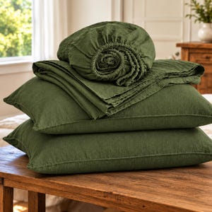 May include: A set of olive green bedding featuring two rectangular pillowcases stacked horizontally, topped with a folded flat sheet and a fitted sheet with visible elastic edges, all made from a textured fabric.