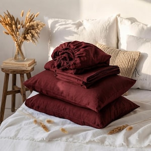May include: A set of deep burgundy linen bedding, including two rectangular pillowcases and a folded fitted sheet, stacked neatly on a white bedspread. The fabric has a visible natural weave texture, showcasing a rich, dark red color throughout.