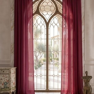 May include: Two floor-length, deep burgundy curtains hang from a silver rod over a tall, arched window with intricate metal lattice detailing. The sheer fabric drapes in vertical folds, framing the view of an outdoor courtyard with trees.