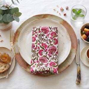May include: A rectangular cloth napkin featuring a repeating floral pattern with vibrant pink flowers and green foliage on a white background, neatly folded and centered on a speckled ceramic dinner plate with a rustic brown rim.