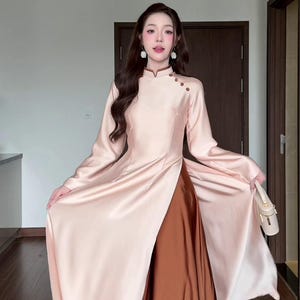 May include: A long-sleeved, floor-length dress featuring a pale pink satin tunic with a high collar, side buttons, and a high side slit, worn over a solid dark brown satin skirt. The garment includes a small beige handbag and white floral earrings.