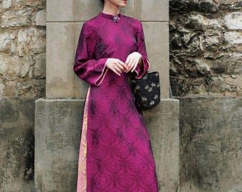 May include: A long-sleeved magenta tunic with a tonal circular pattern, featuring a high collar with a decorative brooch, side slits revealing a light pink panel, and loose-fitting green trousers, paired with a small black patterned handbag.