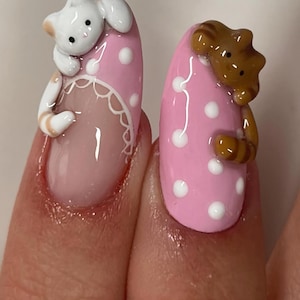 May include: Close-up of two manicured nails. One nail is pink with white polka dots and a white cat figurine. The other nail is pink with white polka dots and a brown cat figurine. The nails have a glossy finish.