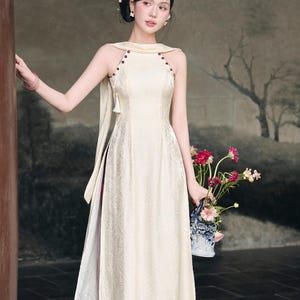 May include: A floor-length cream-coloured sleeveless dress featuring a high halter neckline with dark red button accents, a subtle tonal jacquard pattern, and a long fabric sash draped from the shoulder. The dress has side slits revealing a white layer.
