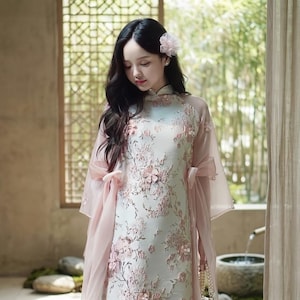 Könnte beinhalten: Ein helles, floral gemustertes Kleid mit Stehkragen und rosa Akzenten. Das Kleid hat 3D-Blütenverzierungen und wird mit einem transparenten rosa Schal kombiniert. Eine weiße Handtasche mit Perlenriemen wird in der Hand gehalten.