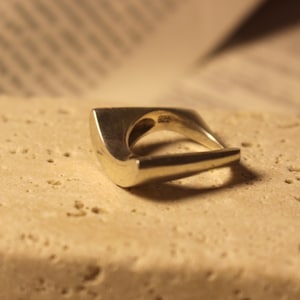 May include: A silver ring with a unique, geometric design. The ring features a curved band that forms a square shape on top, with a pointed end. The ring is set on a textured, light-colored surface.