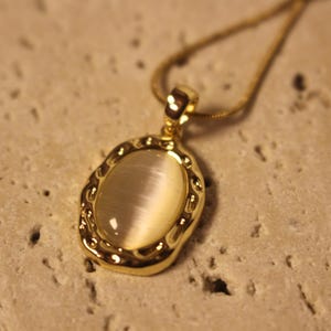 Puede incluir: Un collar dorado con un colgante ovalado con una piedra de color crema. El colgante tiene un marco texturizado y adornado. El collar descansa sobre una superficie texturizada y de color claro.