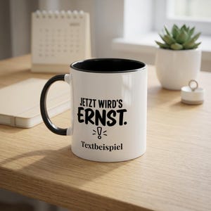 Puede incluir: Una taza de cerámica blanca con interior y asa de color negro. El frente presenta texto negro que dice "JETZT WIRD'S ERNST." con un icono de signo de exclamación debajo, seguido del texto negro más pequeño "Textbeispiel".