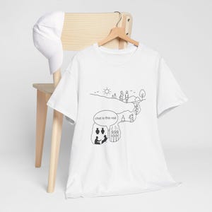 Op de afbeelding: Wit T-shirt met een zwarte lijntekening van een landschap en figuren. De tekst "chat is this real" staat in een tekstballon. Een witte baseballpet hangt aan een houten stoel.