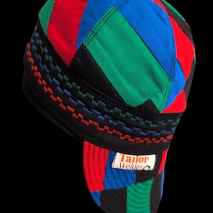 May include: A colorful welding cap with a patchwork design. The cap features red, blue, green, and black panels. The brim has a white label with the text "Tailor Welder". The cap has a black band with decorative stitching.