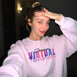 May include: A light pink sweatshirt with the text "Jessica Virtual Assistant" in a colorful design. The sweatshirt has long sleeves and a crew neck. The image is taken indoors.