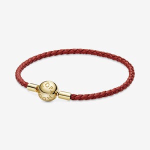 Puede incluir: Una pulsera de cuero trenzado rojo con un cierre dorado. El cierre es una esfera redonda con el nombre de la marca grabado. La pulsera es un accesorio elegante.