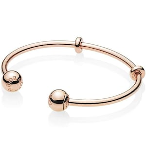 May include: Rose gold open cuff bracelet with spherical end caps and a central accent. The bracelet has a smooth, polished finish and a minimalist design. The spherical end caps have engraved text.