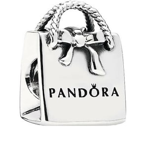 May include: A silver charm in the shape of a shopping bag, featuring a braided handle and a bow detail. The word "PANDORA" is printed in black on the front. This charm is designed for bracelets.