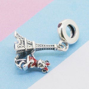 May include: A silver charm featuring the Eiffel Tower and a small figure in a red outfit. The charm has a round, open-ended design for attachment to a bracelet or necklace. The charm is detailed with intricate designs.
