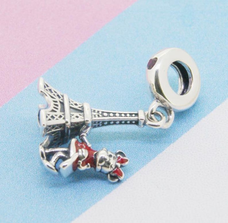 May include: A silver charm featuring the Eiffel Tower and a small figure in a red outfit. The charm has a round, open-ended design for attachment to a bracelet or necklace. The charm is detailed with intricate designs.