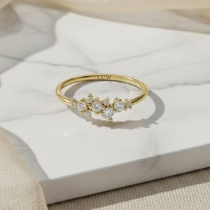 May include: A gold ring featuring a cluster of small, clear, round-cut stones arranged in a delicate, asymmetrical pattern along the top of the band. The interior of the gold band is clearly engraved with the letters LUW in a simple, clean font.