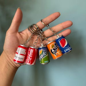 Pode incluir: Um conjunto de cinco porta-chaves de latas de bebida em miniatura presos a um anel de metal prateado. A coleção inclui réplicas de marcas como Budweiser, Coca-Cola, Sprite, Fanta Orange e Pepsi, cada uma com o seu logótipo e design.