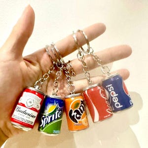 May include: A collection of miniature soda can keyrings, featuring brands like Budweiser, Sprite, Fanta, Coca-Cola, and Pepsi. Each keyring showcases a detailed miniature replica of the can design, attached to a silver chain. A novelty item for fans of fizzy drinks.