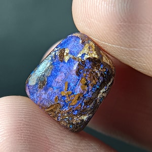 Könnte beinhalten: Ein kleiner, polierter Boulder-Opal-Edelstein in rechteckiger Form. Die Oberfläche zeigt ein lebhaftes, irisierendes blaues Farbspiel, das mit dunklen, erdig braunen Eisenstein-Matrix-Einschlüssen auf der texturierten Vorderseite kontrastiert.
