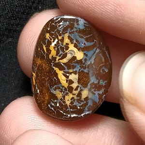 Könnte beinhalten: Ein polierter, oval geformter Opal-Edelstein. Der Stein hat eine dunkelbraune Grundfarbe mit komplizierten Mustern aus Gelb, Blau und Weiß, die ein einzigartiges, natürliches Design ergeben. Der Opal wird zwischen Fingern gehalten.