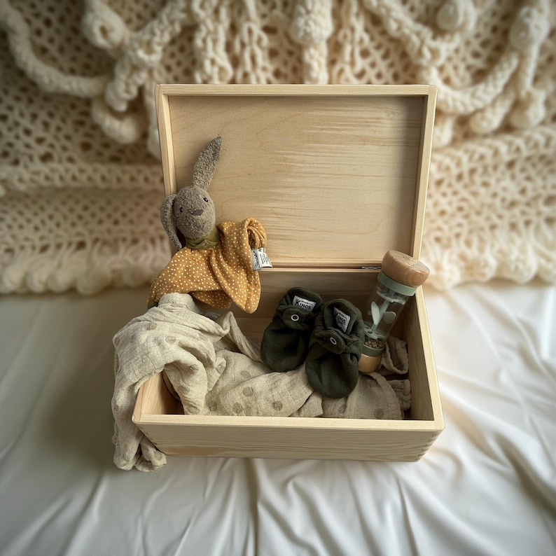 May include: A wooden box with a hinged lid, filled with baby items. Includes a stuffed bunny, a yellow and white polka dot blanket, olive green baby shoes, a muslin blanket, and a clear bottle with a wooden top.