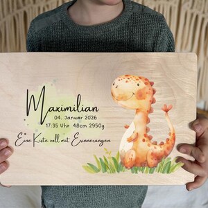 May include: A wooden plaque with the name "Maximilian" in black script, along with birth details and a watercolor illustration of a friendly orange dinosaur. The text "Eine Kiste voll mit Erinnerungen" is also included.