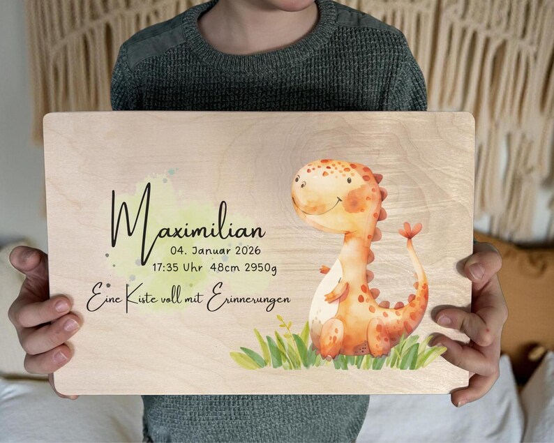 May include: A wooden plaque with the name "Maximilian" in black script, along with birth details and a watercolor illustration of a friendly orange dinosaur. The text "Eine Kiste voll mit Erinnerungen" is also included.