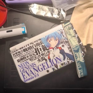 May include: A white and blue wallet featuring an anime character with blue hair and the text "NEON GENESIS EVANGELION" and Japanese text. The wallet has a matching wrist strap with a character image.