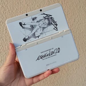 May include: A light blue Nintendo 3DS handheld game console, open to reveal a screen with a black and white anime illustration. The text "GROUNDWORK OF EVANGELION 2.0" is printed on the bottom half of the console.