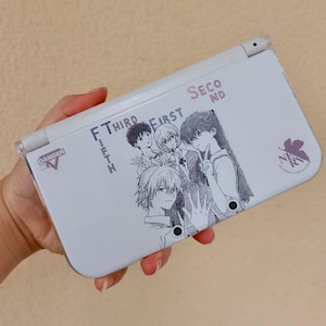Könnte beinhalten: Eine weiße Handheld-Spielkonsole mit einer Schwarz-Weiß-Anime-Illustration von vier Charakteren. Die Konsole trägt die Schriftzüge "SECOND", "THIRD", "FIRST" und "FIFTH". Die Konsole trägt auch die Schriftzüge "DANGER" und "TV".